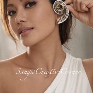 May include: An ornate ear cuff featuring a spiral design with a central black stone, surrounded by pearls and colorful gemstones. The cuff is held near the ear, showcasing its intricate details and craftsmanship. A delicate gold necklace is also visible.
