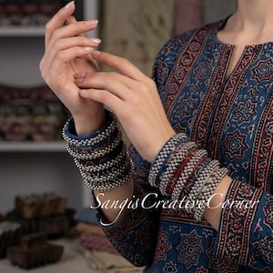 May include: A person wearing multiple beaded bracelets in shades of blue, red, and silver. The bracelets are stacked on the wrist. The person is wearing a blue and red patterned top. The text "Sangis Creative Corner" is visible.