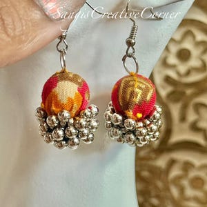 May include: A pair of handmade earrings. Each earring features a red and gold patterned fabric ball, topped with silver-colored metal beads. The earrings hang from silver-colored fish hook ear wires. The text "Sandis Creative Corner" is visible.
