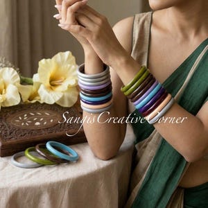 May include: A collection of smooth, solid-colored bangle bracelets in shades of gray, lavender, brown, blue, and green. The bracelets are stacked on both wrists, with additional loose bangles resting on a wooden box. Text reads Sangis Creative Corner.