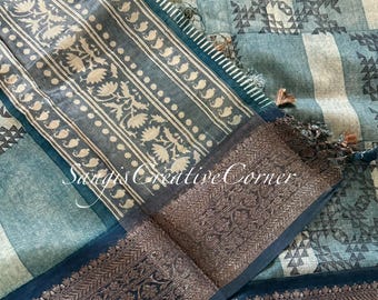 May include: A teal blue and white cotton saree with a geometric pattern and a gold and black border. The saree has a fringe trim and a small tassel.