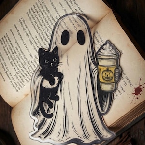 May include: A flat, die-cut acrylic bookmark featuring a white ghost with black eyes. The ghost holds a black cat in one arm and a beige coffee cup with a yellow sleeve displaying a jack-o'-lantern face in the other hand. The design has black outlines.