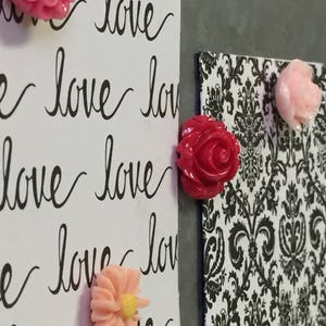 May include: A close-up of a white board with the word "love" written on it multiple times in black cursive. There are four pink and red flower-shaped magnets attached to the board.