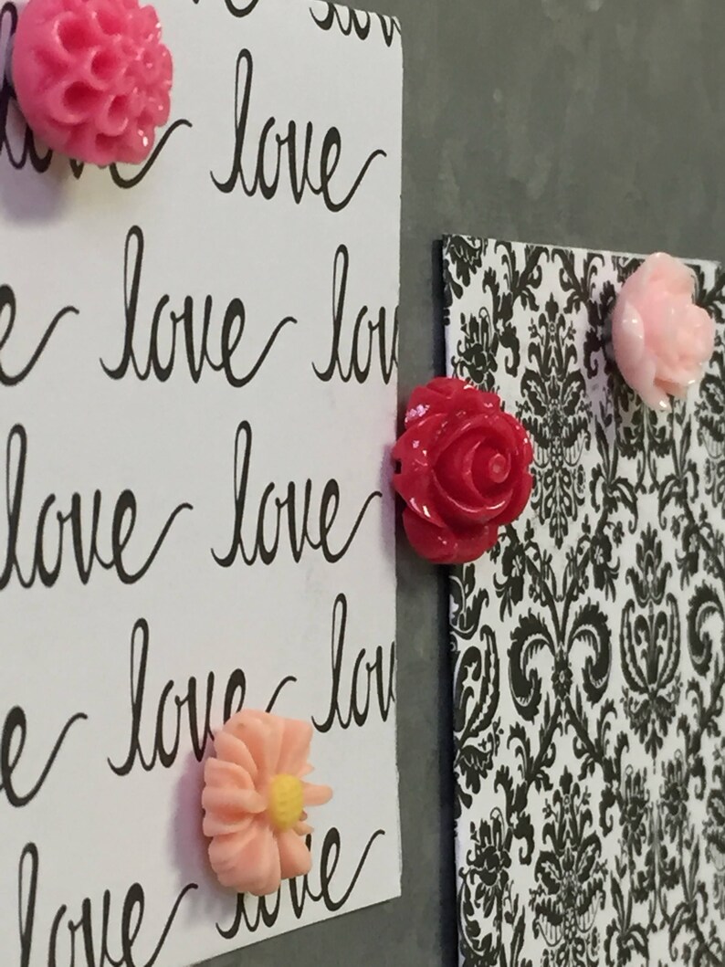 May include: A close-up of a white board with the word "love" written on it multiple times in black cursive. There are four pink and red flower-shaped magnets attached to the board.