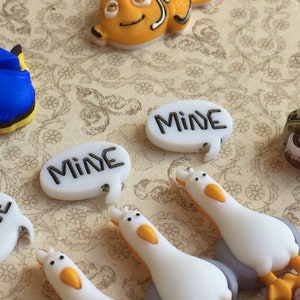 May include: A collection of colorful resin embellishments featuring characters from a popular animated film. Includes a blue fish, an orange fish, a green turtle, and white seagulls. Several speech bubbles say "MINE" in black lettering.