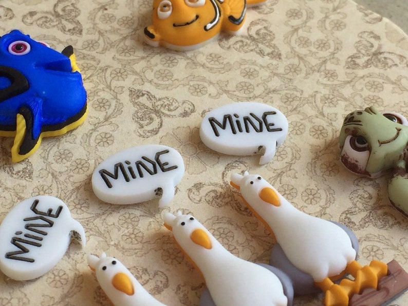 May include: A collection of colorful resin embellishments featuring characters from a popular animated film. Includes a blue fish, an orange fish, a green turtle, and white seagulls. Several speech bubbles say "MINE" in black lettering.