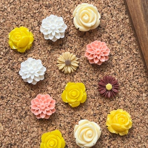 Puede incluir: Surtido de 11 chinchetas con forma de flor de colores en varios tonos de amarillo, blanco, rosa y marrón. Las chinchetas están dispuestas sobre un fondo de corcho.