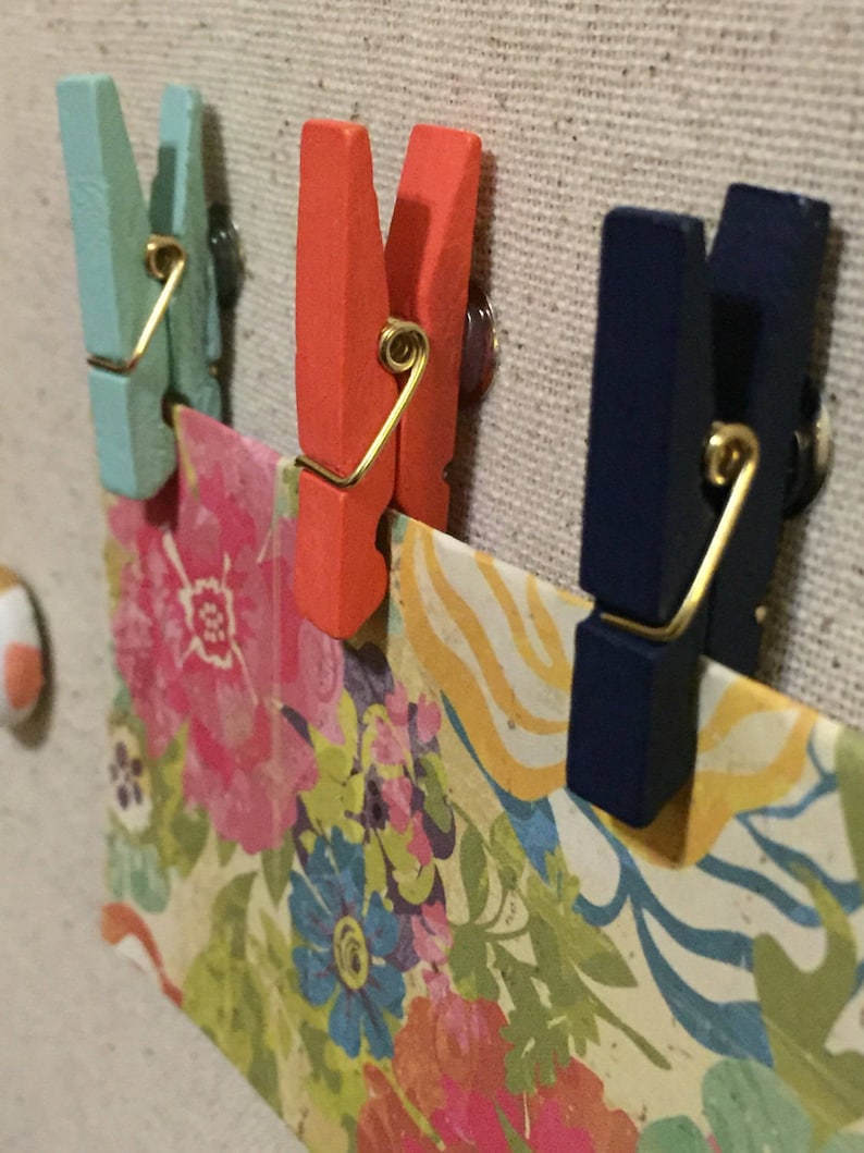 May include: Three wooden clothes pegs with metal springs. The clothes pegs are painted in different colours: teal, orange, and navy blue. The clothes pegs are holding a piece of patterned paper with flowers and swirls.