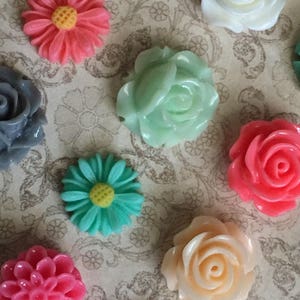 May include: A collection of colorful resin flower embellishments in various shades of pink, green, white, and peach. The flowers include roses and daisies, with yellow centers. The background is a patterned, neutral-toned fabric.