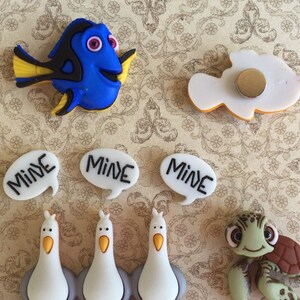 May include: A collection of colorful, cartoon-style magnets featuring characters from the movie "Finding Nemo." Includes a blue fish, a white fish, seagulls, a sea turtle, and speech bubbles with the word "MINE."