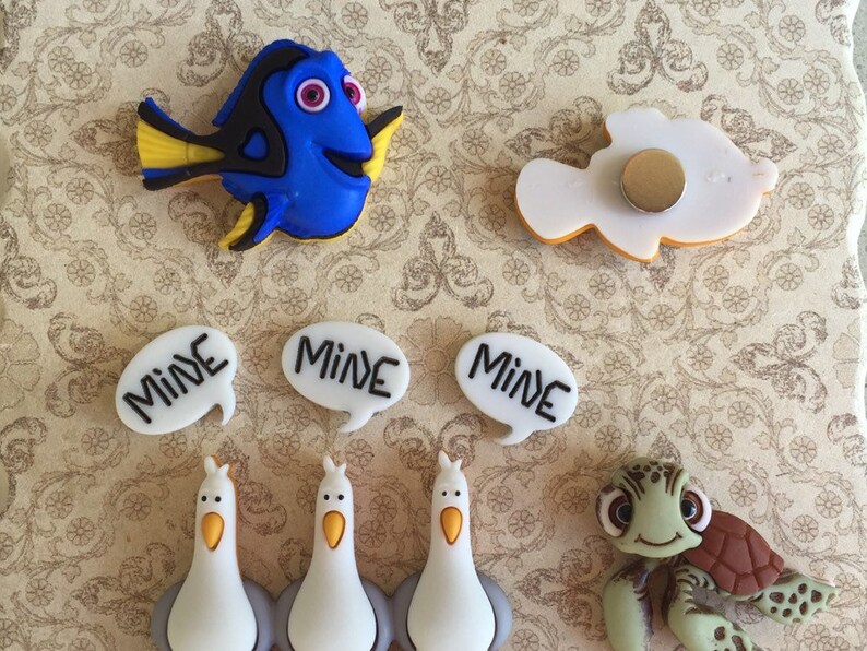 May include: A collection of colorful, cartoon-style magnets featuring characters from the movie "Finding Nemo." Includes a blue fish, a white fish, seagulls, a sea turtle, and speech bubbles with the word "MINE."