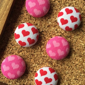 May include: Six fabric-covered push pins with heart patterns. Three pins have a white background with red hearts and three pins have a pink background with white hearts.