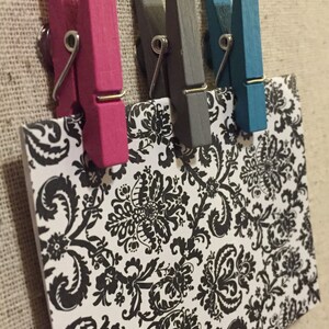 May include: Four wooden pegs in different colours: pink, grey, blue, and teal. The pegs are holding a card with a black and white damask pattern.