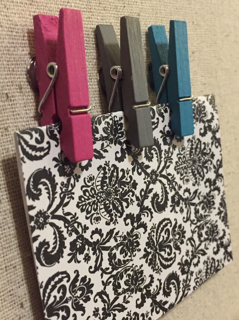 May include: Four wooden pegs in different colours: pink, grey, blue, and teal. The pegs are holding a card with a black and white damask pattern.