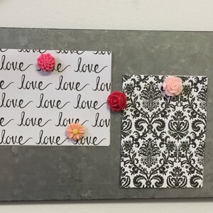 May include: A gray metal board with two white cards. One card has the word "love" written repeatedly in black cursive. The other card has a black and white damask pattern. There are four pink and orange flower-shaped push pins on the cards.
