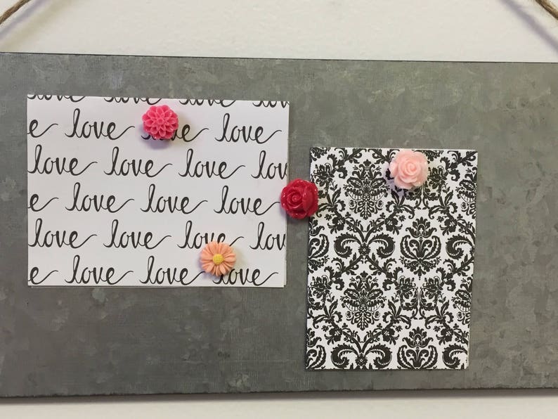 May include: A gray metal board with two white cards. One card has the word "love" written repeatedly in black cursive. The other card has a black and white damask pattern. There are four pink and orange flower-shaped push pins on the cards.