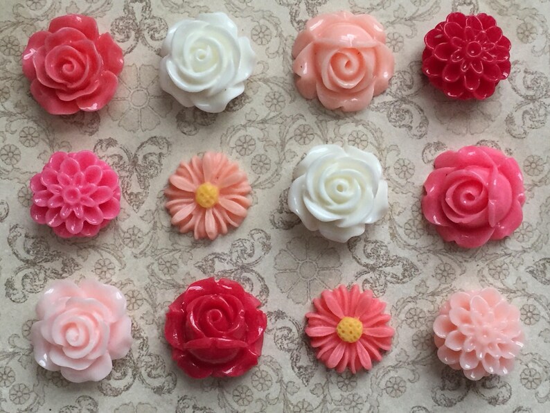 May include: Twelve assorted resin flower embellishments in shades of pink, white, and red. The flowers are in various styles, including roses, daisies, and carnations.