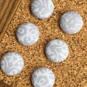 May include: Six grey fabric push pins with a white floral pattern on a cork board.