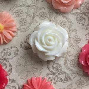 May include: Assortment of white, pink, and red plastic flower embellishments. The flowers are in various shapes and sizes, including roses and daisies.