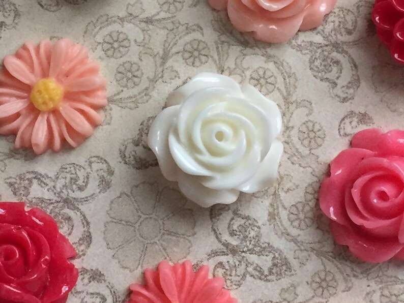 May include: Assortment of white, pink, and red plastic flower embellishments. The flowers are in various shapes and sizes, including roses and daisies.