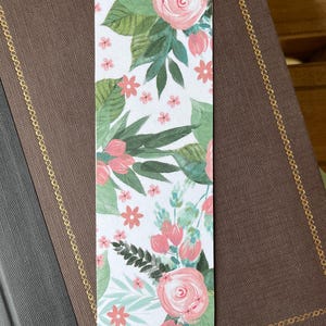 May include: A floral bookmark featuring a watercolor design of pink roses, small pink flowers, and green leaves on a white background. The bookmark is rectangular with rounded corners and is resting on a brown book.