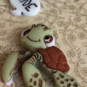 May include: A green turtle figurine with a brown shell and a smiling face. The turtle has light brown spots on its flippers and legs. A white speech bubble with the word "MINE" in black is also present.