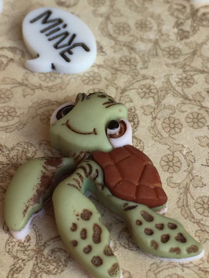 May include: A green turtle figurine with a brown shell and a smiling face. The turtle has light brown spots on its flippers and legs. A white speech bubble with the word "MINE" in black is also present.