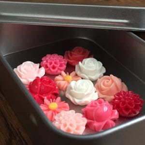 May include: A tin box filled with a variety of pink, red, and white flower-shaped buttons. The buttons are arranged in a single layer in the box.
