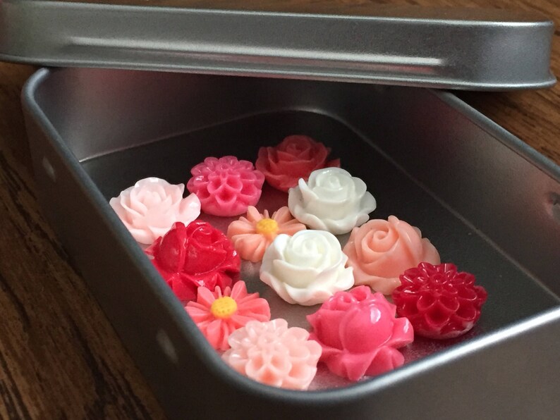 May include: A tin box filled with a variety of pink, red, and white flower-shaped buttons. The buttons are arranged in a single layer in the box.