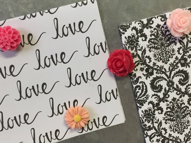 May include: Two white cards with the word "love" written on them in black cursive. The cards are decorated with pink and red flower-shaped push pins.