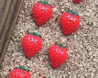 May include: Nine red strawberry-shaped push pins with green leaves on a corkboard background.