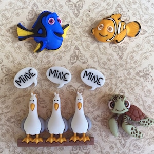 May include: A collection of colorful character-themed embellishments. Includes a blue fish, an orange fish, three white speech bubbles with "MINE" text, three seagulls, and a green turtle. These are likely for crafting or scrapbooking.