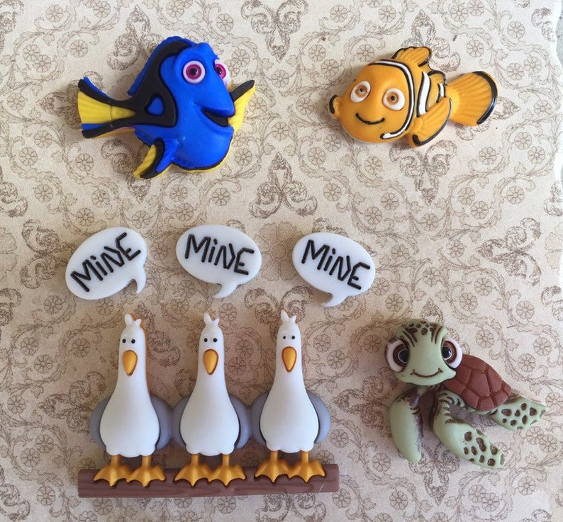 May include: A collection of colorful character-themed embellishments. Includes a blue fish, an orange fish, three white speech bubbles with "MINE" text, three seagulls, and a green turtle. These are likely for crafting or scrapbooking.