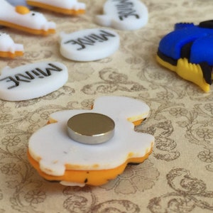 May include: A collection of colorful magnets featuring cartoon characters and the word "MINE". The magnets include a blue and yellow fish, and white speech bubbles with black text. The magnets have a flat back with a silver magnet attached.