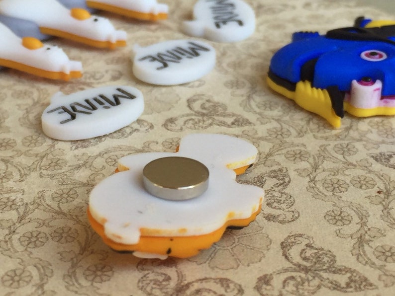 May include: A collection of colorful magnets featuring cartoon characters and the word "MINE". The magnets include a blue and yellow fish, and white speech bubbles with black text. The magnets have a flat back with a silver magnet attached.