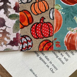 Pode incluir: Marcadores artesanais com temas de outono. Os designs incluem folhas de outono, abóboras com bolinhas e listras e uma ilustração de abóbora em um fundo turquesa. Os marcadores são colocados em cima de um livro aberto.