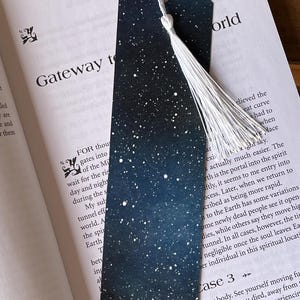 May include: A dark blue bookmark with white stars printed on it. The bookmark has a white tassel attached to it. The bookmark is laying on a page of a book with text about a gateway to another world.