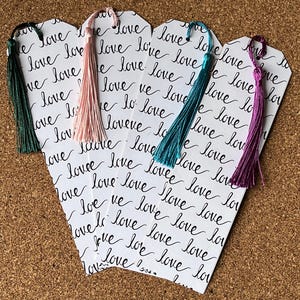 Puede incluir: Cinco marcapáginas blancos con escritura cursiva negra que dice "love" repetidamente. Cada marcapáginas tiene un borla unido a la parte superior. Las borlas son de color rosa, turquesa, morado y dos tonos de verde.