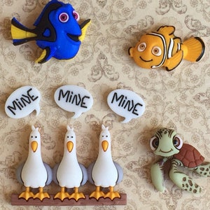 May include: A collection of cartoon character embellishments, including a blue fish, an orange fish, three white seagulls, a green turtle, and three speech bubbles with the word "MINE". The embellishments are on a patterned background.