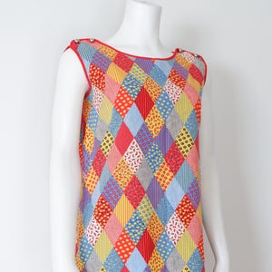 May include: A sleeveless, knee-length dress with a patchwork design. The dress features a vibrant pattern of red, yellow, blue, and white diamond shapes. The neckline and armholes are trimmed in red. Silver buttons adorn the shoulders.