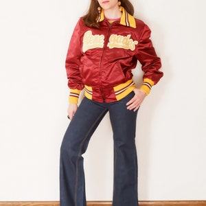 May include: A red satin bomber jacket with yellow and white striped trim. The jacket has "Renegades" written in yellow on the front. The model is wearing blue jeans and colorful polka dot shoes.