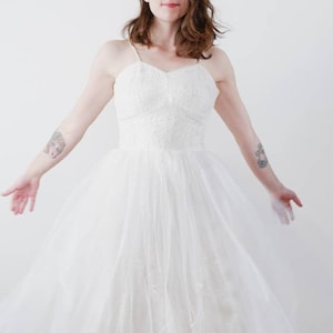 May include: A white tulle wedding dress with a lace bodice and a sweetheart neckline. The dress has a full skirt and is worn with white shoes.