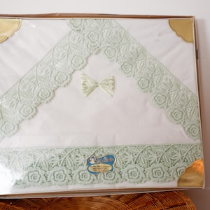 May include: A white lace bed sheet with green lace trim and a green satin bow in the centre. The sheet is packaged in a clear plastic bag with a gold label that reads "Baby's First Bed Sheet Set".