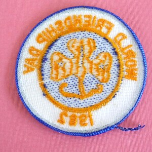 Puede incluir: Un parche redondo con borde azul y reverso blanco. El parche presenta un emblema naranja con las palabras "Friendship Day" y "1985" bordadas en naranja. El emblema est&aacute; centrado en un fondo azul claro punteado.