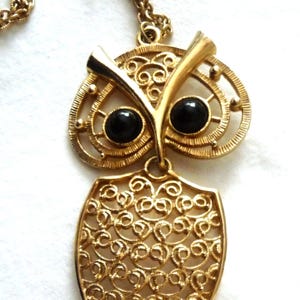 May include: Gold-toned owl pendant necklace. The owl pendant features a detailed filigree design with black circular eyes. The necklace chain is also gold-toned. The owl's body is a shield shape with a detailed openwork design.
