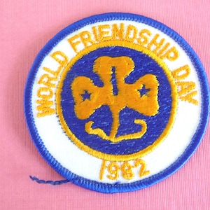 Puede incluir: Un parche redondo con borde azul y centro blanco. El parche presenta texto dorado que dice "WORLD FRIENDSHIP DAY" y "1982". En el interior hay un dise&ntilde;o dorado en forma de tr&eacute;bol con dos estrellas.