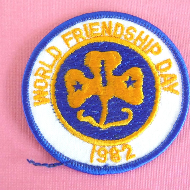 Puede incluir: Un parche redondo con borde azul y centro blanco. El parche presenta texto dorado que dice "WORLD FRIENDSHIP DAY" y "1982". En el interior hay un dise&ntilde;o dorado en forma de tr&eacute;bol con dos estrellas.