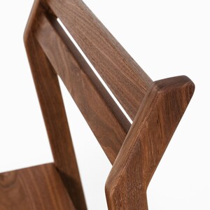 May include: Close-up of a wooden chair back. The chair is made of dark brown wood with a smooth finish. The design features a slatted backrest and angled supports, showcasing a modern aesthetic.