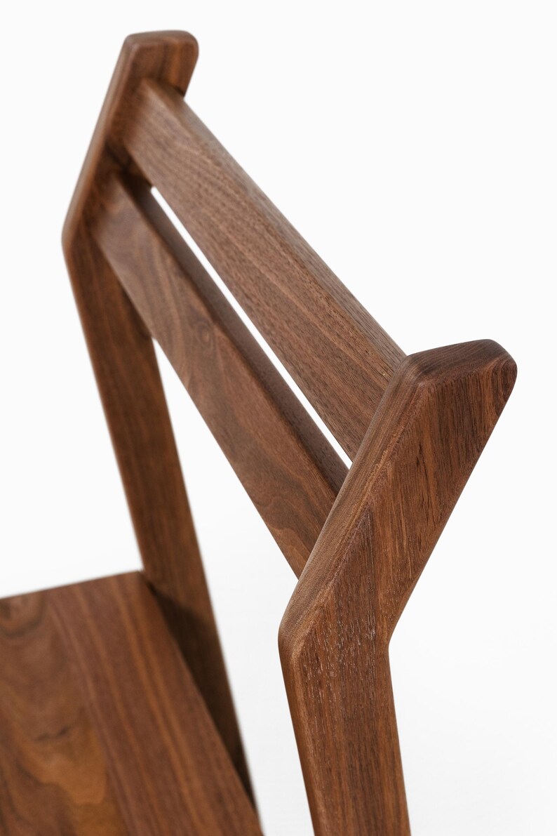May include: Close-up of a wooden chair back. The chair is made of dark brown wood with a smooth finish. The design features a slatted backrest and angled supports, showcasing a modern aesthetic.