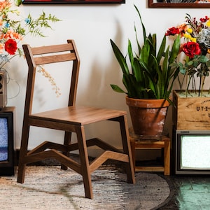 May include: A wooden chair with a unique angled leg design stands in front of a wall. Two vintage televisions with static screens are on the floor. Colorful flowers are arranged in vases and wooden crates, adding a vibrant touch to the scene.